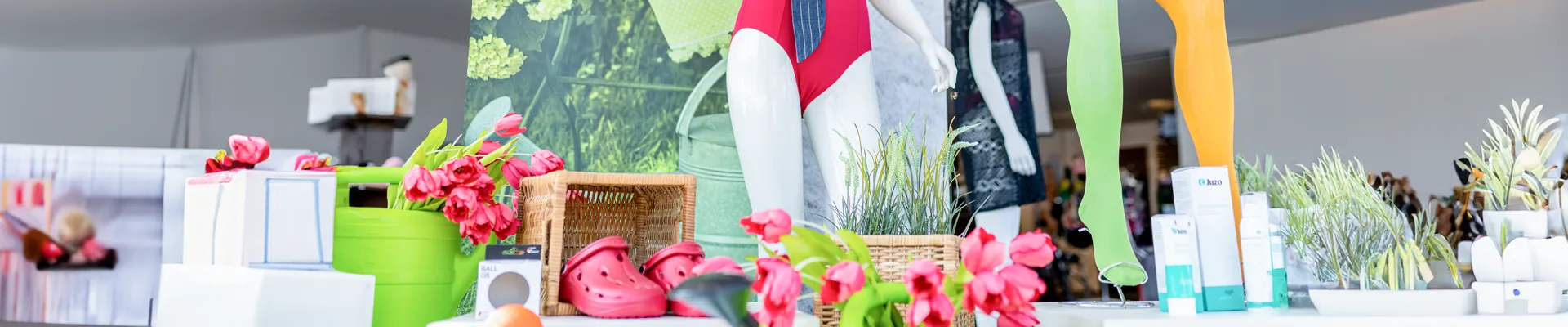 es sind Sanitätsartikel zu sehen. rechts auf dem Bild sind ein gelber und ein grüner Kompressionsstrumpf zu sehen. links daneben ist ein Teil von einem roten Badeanzug zu sehen. Leicht links davor sind rote Hausschuhe zu sehen. Links daneben befindet sich eine grüne Gießkanne.