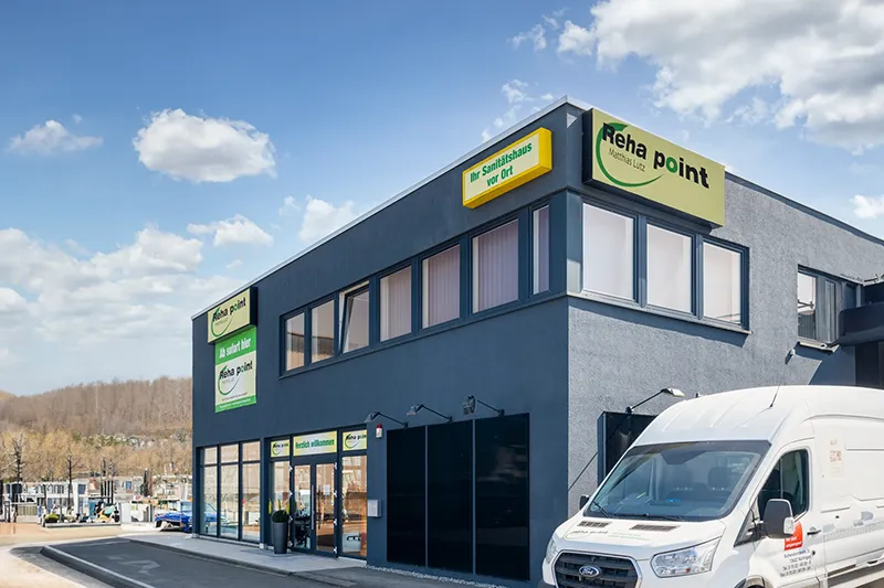 Es ist das Gebäude von Reha Point in Nürtingen zu sehen. Es ist ein graues Gebäude mit mit gelber Reklame mit der Werbung von Reha Point Im Vordergrund steht ein weißer Transporter von Reha Point.