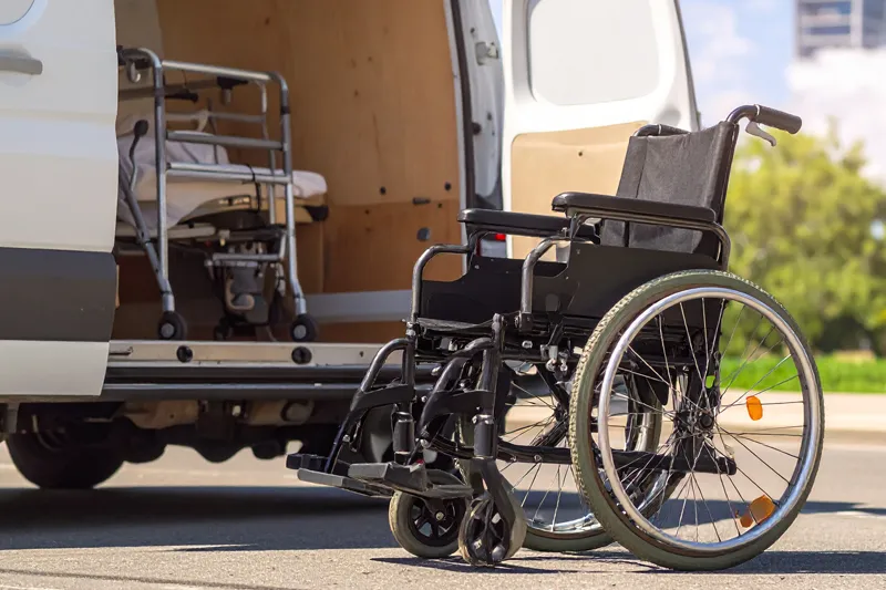 Ein Rollstuhl steht leer auf der Straße vor einem Kastenwagen. Im Kastenwagen sieht man im Ansatz ein Pflegebett