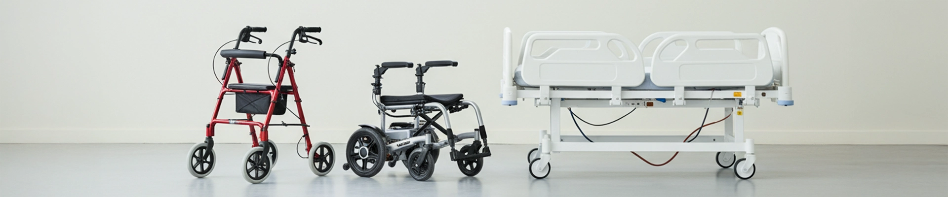 Vor einer weißen Wand stehen 3 unterschiedliche Hilfsartikel für den Reha Bedarf. Ganz links steht ein roter Rollator. In der Mitte steht ein elektrischer Rollstuhl und ganz links ist ein elektrisches Pflegebett zu erkennen. Beim Bett sind seitlich Herausfallsicherungen angebracht.