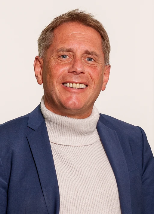 Porträtfoto bis zur Brust von Matthias Lutz vor einer weißen Wand.Er trägt einen beigefarbenen Rollkragenpullover und darüber ein blaues Sakko.<br />